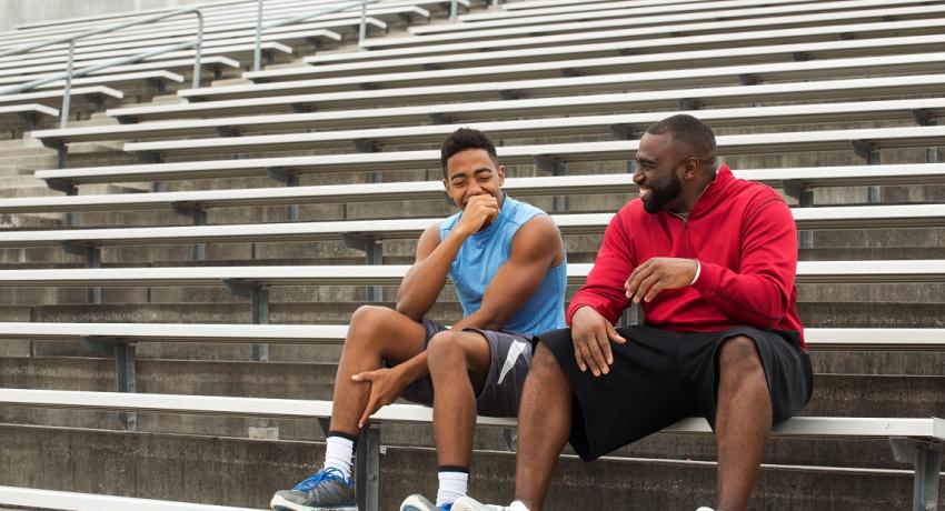 coach sitting with student athlete on bleachers in stadium, both laughing. 