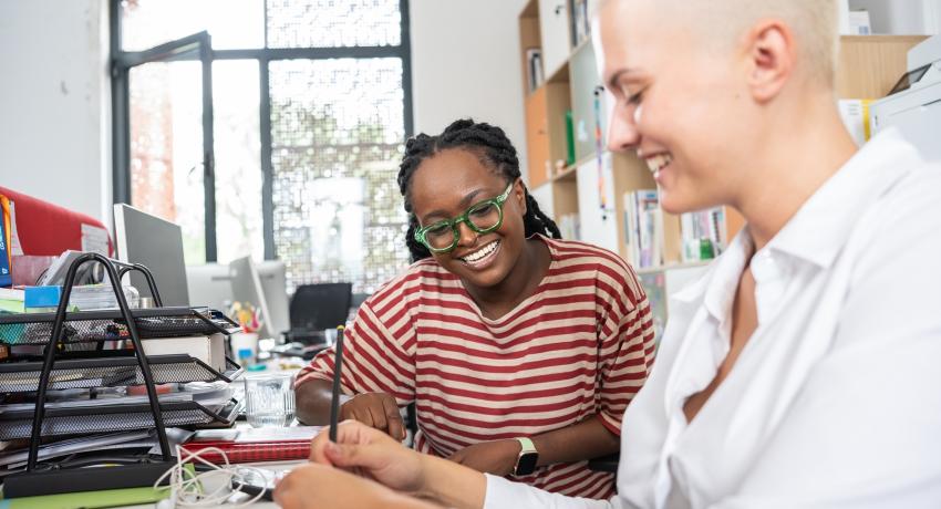 Two coworkers sitting at a desk looking at a shared document and smiling. 