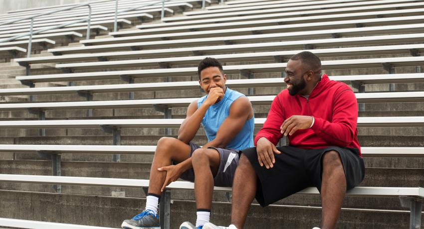 A teenager and his mentor or coach sit on bleachers in a stadium talking and smiling. 