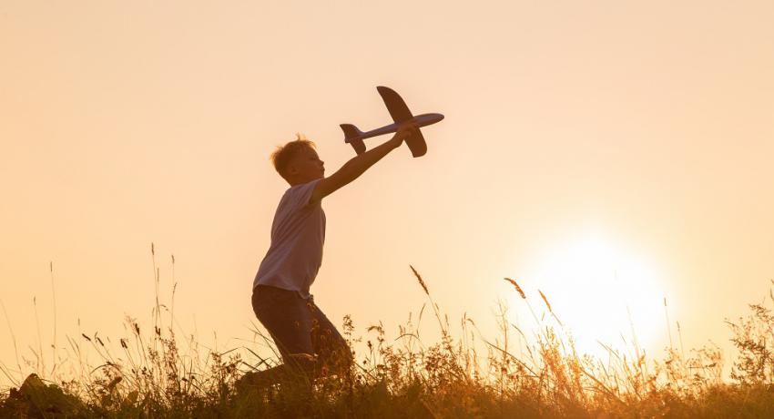 Kid playing in a meadow with big toy plane during gold sunset in summer landscape.