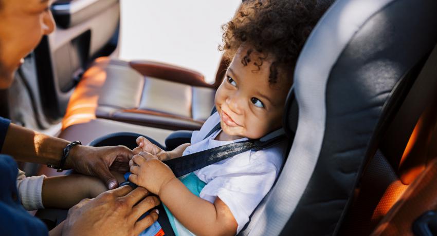 A smiling caregiver buckles a toddler in a car seat. 