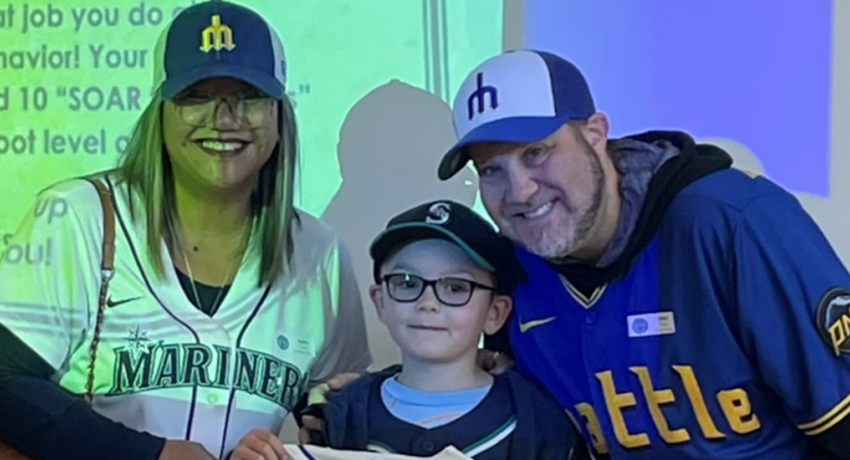 Photo of two adults with a child in the middle smiling at the camera dressed in Seattle Mariners Baseball gear