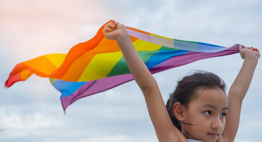 Young girl with dark hair holding up high an unfurled, colorful Pride Flag.