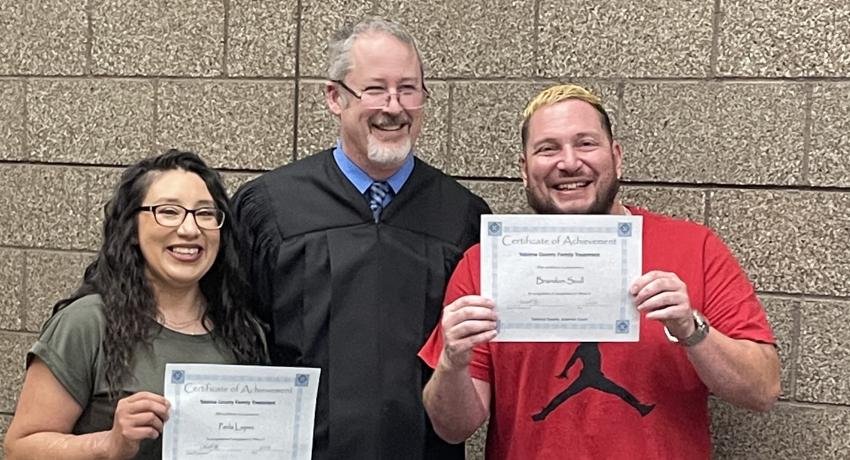 Two parents smiling holding up their certificates on either side of a judge