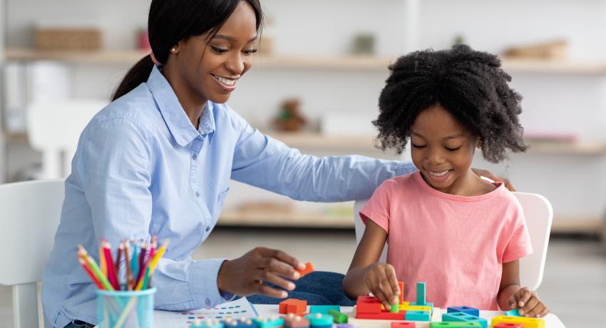 Woman of color sits with girl of color in early learning classroom.