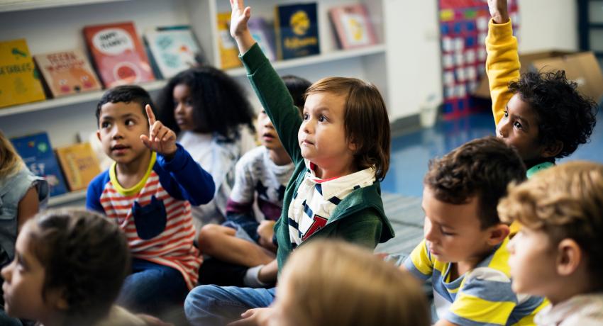 Young children sitting in class on carpet raising hands. 