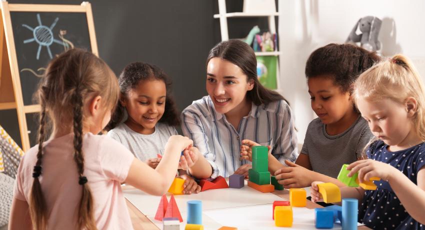 Woman at table in classroom talking and playing with four children.