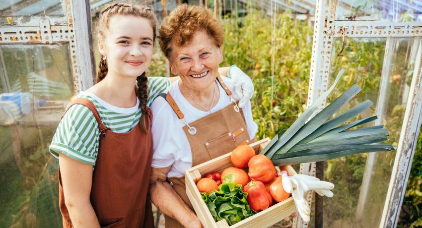 granddaughter and grandma with box of veggies smiling outside a greenhouse